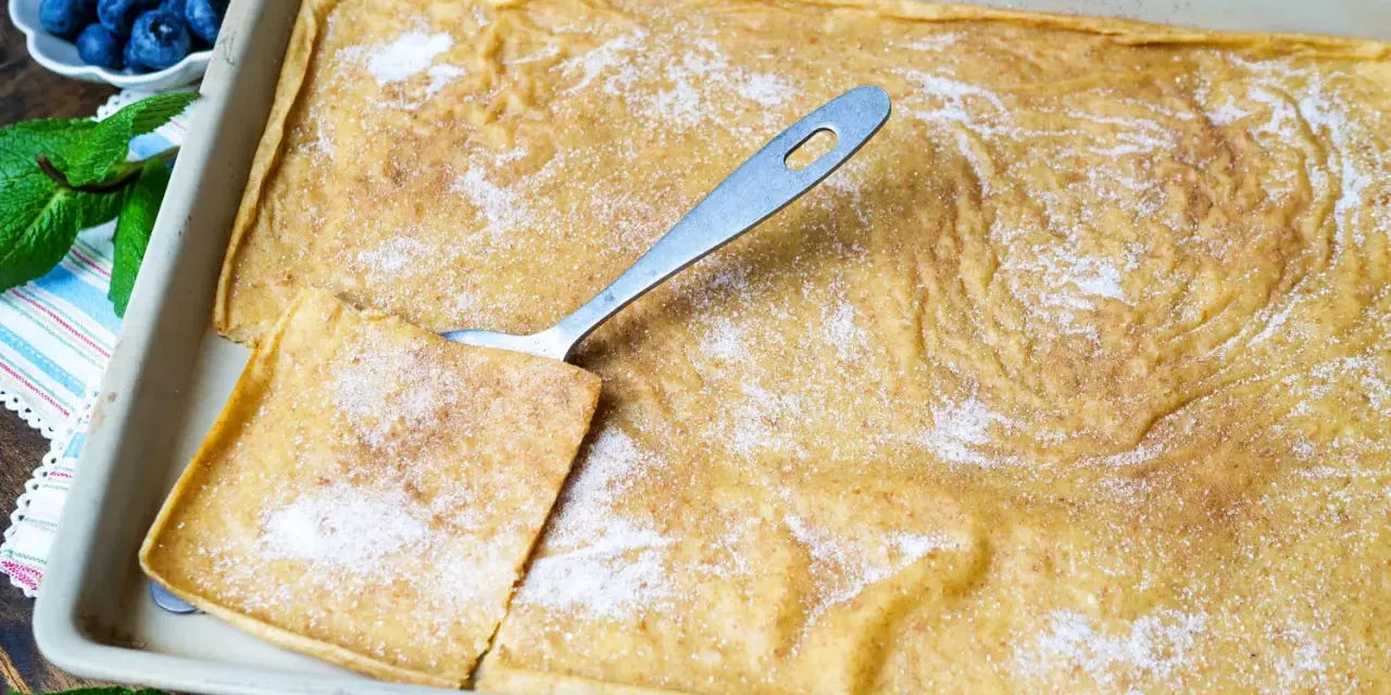 Ein frisch gebackener Ofenpfannkuchen, bestreut mit Zucker und Zimt, bereit zum Servieren. Ein Stück wird mit einem Löffel herausgehoben.