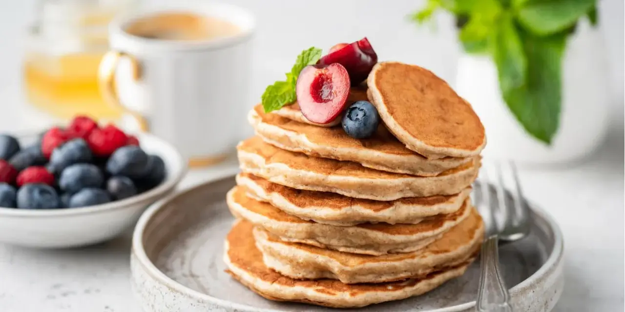 Ein Stapel veganer Pancakes, garniert mit Kirschen und Blaubeeren, daneben eine Schale Beeren und eine Tasse Kaffee.