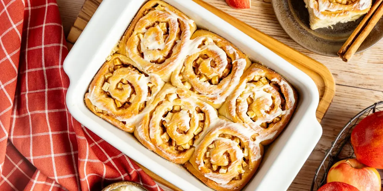 Frisch gebackene Apfel Zimt Schnecken mit Zuckerguss in einer Auflaufform. Daneben Äpfel und Zimtstangen.