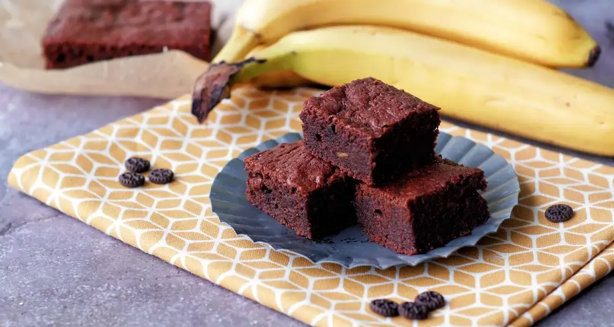 Saftige Bananen Brownies auf einem gelben Tuch, daneben reife Bananen. Ein Genuss!