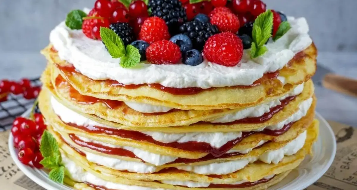 Eine hohe Pfannkuchen Torte mit Sahne und roter Marmelade, gekrönt mit frischen Beeren und Minze.