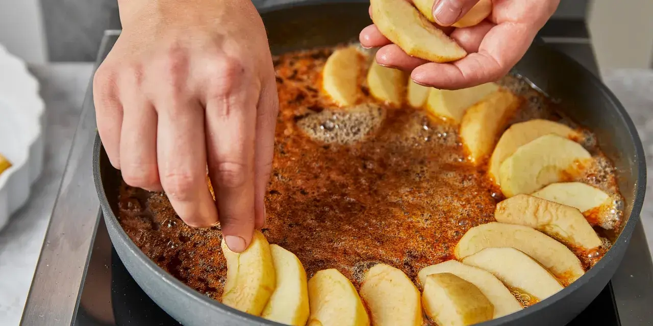 Hände legen Apfelspalten in eine Pfanne mit karamellisierter Butter. Die Zubereitung der klassischen Tarte Tatin beginnt.