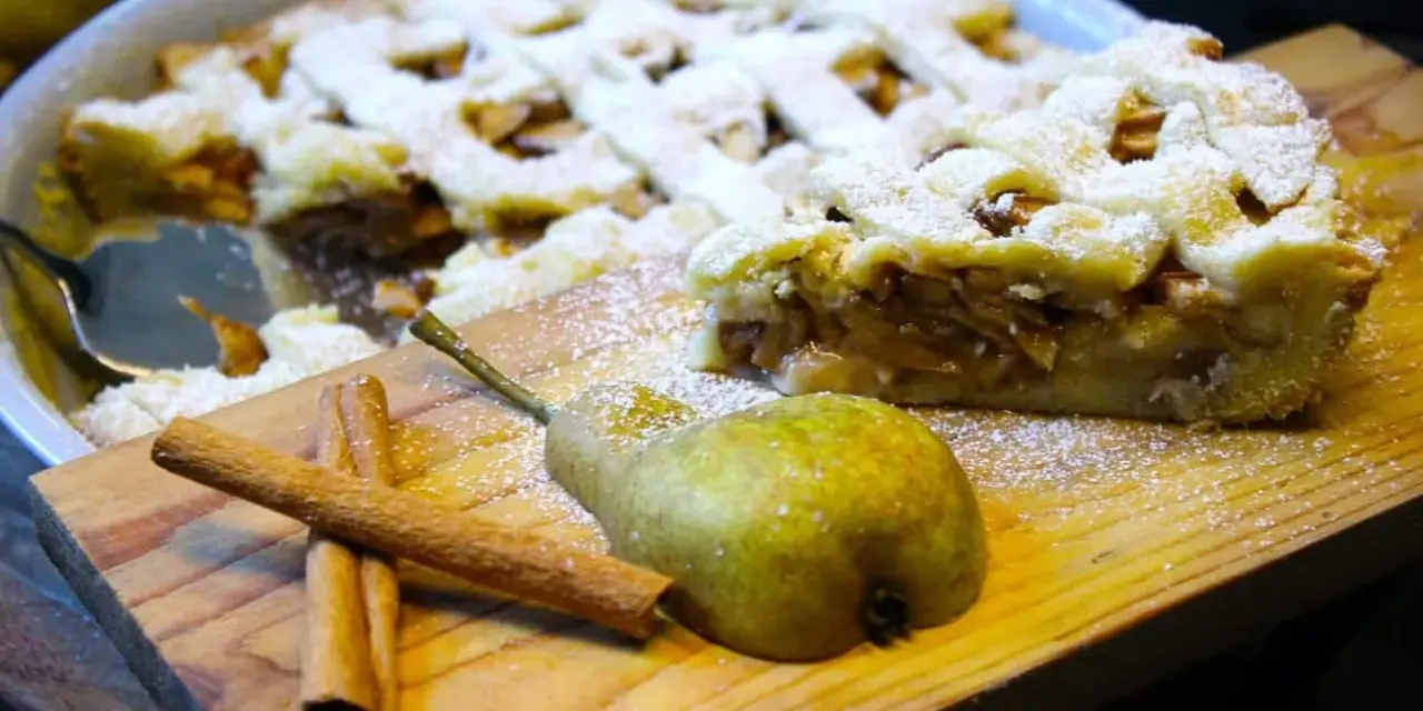 Ein köstlicher Birnenkuchen mit Gittermuster, bestäubt mit Puderzucker. Daneben liegen eine Birne und Zimtstangen – perfekt für ein Birnenkuchen Rezept.