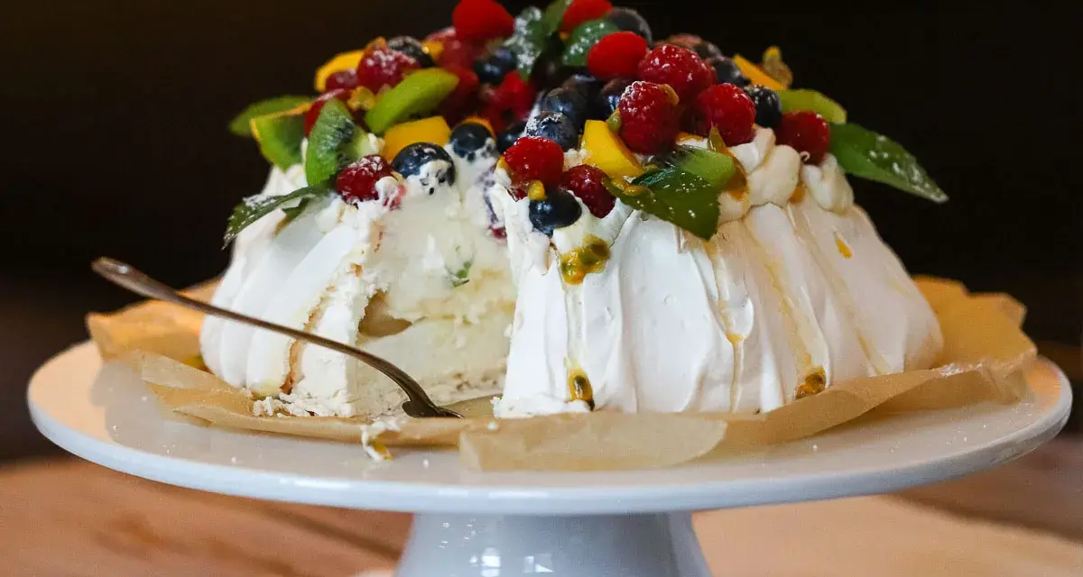Ein köstliches Pavlova-Rezept mit frischen Beeren, Kiwi und Passionsfrucht. Ein Stück ist bereits angeschnitten.