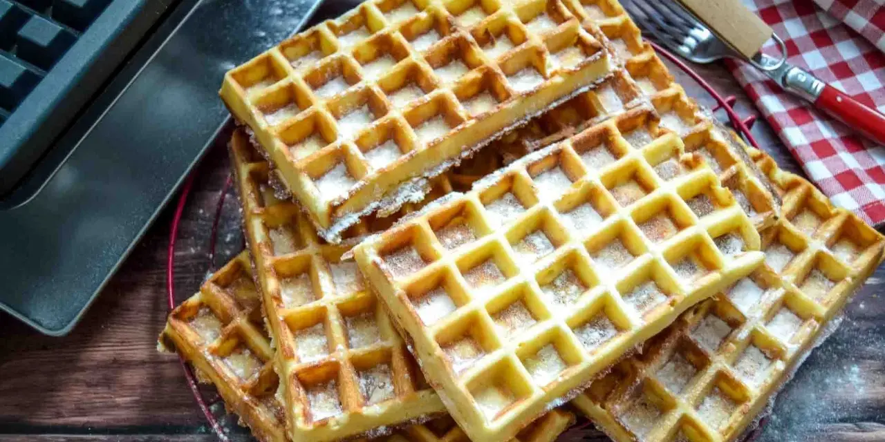 Frisch gebackene belgische Waffeln, mit Puderzucker bestäubt, liegen auf einem Gitter. Daneben ein Waffeleisen und eine karierte Serviette.