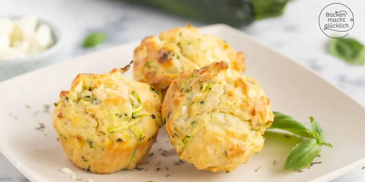 Drei goldbraune Zucchini-Muffins mit Kräutern und Käse auf einem Teller, garniert mit Basilikum.