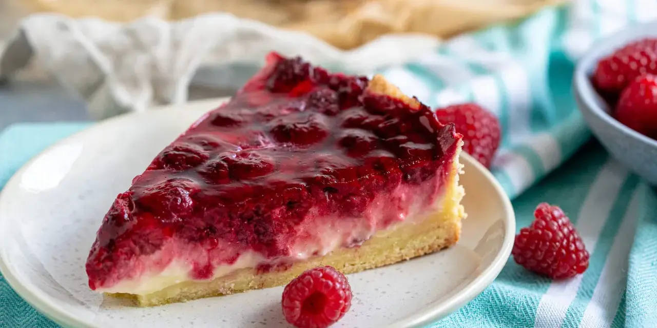 Ein saftiges Stück Himbeer Schmand Kuchen mit frischen Himbeeren und glänzender Glasur auf einem Teller.