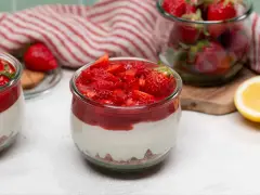 Zwei Gläser mit cremigem Erdbeer-Cheesecake, garniert mit frischen Erdbeeren und Erdbeersauce. Daneben ein Glas voller Erdbeeren und eine Zitronenhälfte.