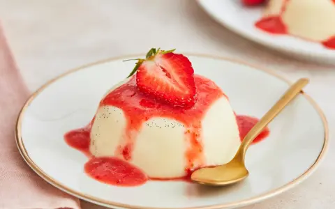 Cremige Panna Cotta mit Erdbeersauce und frischer Erdbeere, garniert mit Vanillepunkten. Eine goldene Löffel liegt daneben.