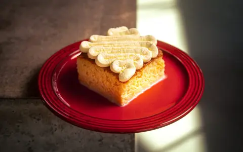 Ein Stück saftiger Tres Leches Kuchen mit Sahnetupfern auf einem roten Teller, bereit zum Genießen.