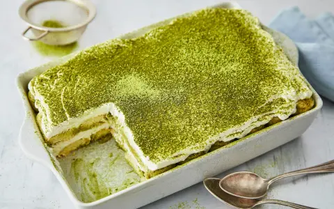 Ein cremiges Matcha Tiramisu, bestäubt mit feinem grünen Pulver, wartet in einer Auflaufform darauf, genossen zu werden. Löffel liegen bereit.