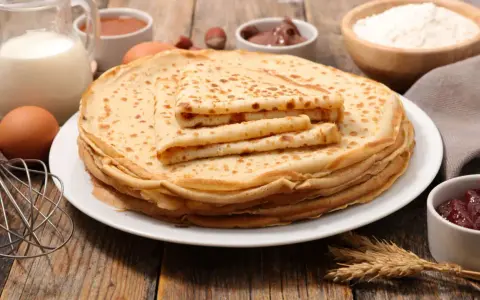 Ein Stapel goldbrauner Pfannkuchen, zubereitet nach einem sehr guten Pfannkuchen Rezept, daneben Zutaten wie Milch, Eier, Mehl, Marmelade und Nuss-Nougat-Creme.