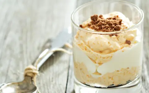 Cremiger cheesecake im glas, geschichtet mit weißer Creme und garniert mit zerbröselten Keksen. Ein Löffel liegt daneben.