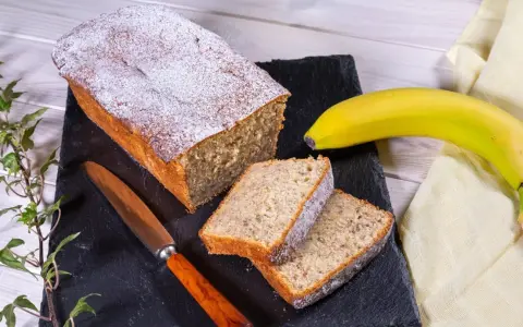 Saftiger Bananenkuchen, bestäubt mit Puderzucker, neben einer Banane und einem Messer. Perfekt für ein Bananenkuchen Rezept.