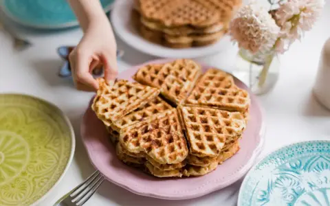 Ein Stapel herzförmiger Waffeln ohne Ei auf einem rosa Teller, bereit zum Genießen. Daneben liegen bunte Teller und eine Gabel.