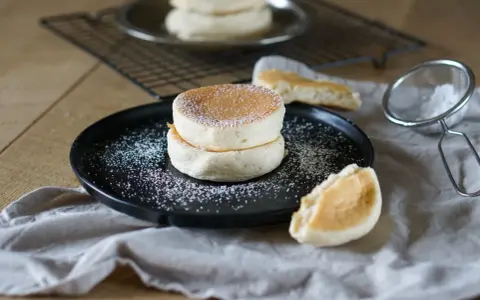 Zwei fluffige japanische Pancakes auf einem schwarzen Teller, bestäubt mit Puderzucker. Daneben ein Stück Pancake und ein Sieb.