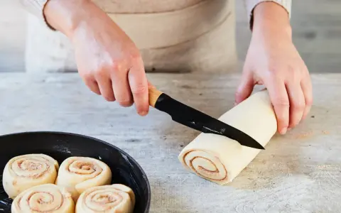 Hände schneiden Teigrolle für Zimtschnecken. Bereits geformte Schnecken warten in einer Pfanne auf das Backen. Ein tolles Zimtschnecken Rezept!