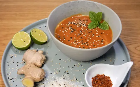 Schüssel mit cremiger Erdnusssauce, garniert mit Sesam und Minze. Daneben Limetten, Ingwer und Chiliflocken – Zutaten für ein leckeres Erdnusssauce Rezept.