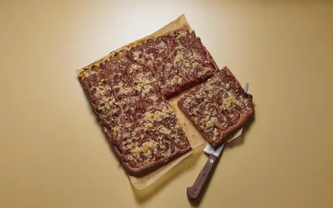 Ein rechteckiger Zwetschgenkuchen mit Streuseln, ein Stück wird mit einem Kuchenheber herausgehoben.