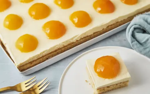 Ein rechteckiger Kuchen mit einer cremigen Schicht und vielen Pfirsichhälften, die wie ein Spiegelei Kuchen aussehen. Ein Stück auf einem Teller daneben.