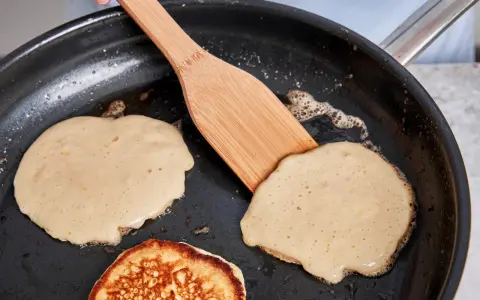 Buttermilch pancakes braten in der Pfanne. Ein Holzspatel wendet einen goldbraunen Pancake.