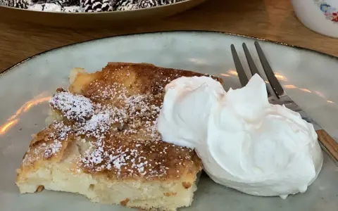 Ein Stück saftiger Apfelkuchen, bestäubt mit Puderzucker, serviert mit einer großen Portion Schlagsahne und einer Gabel.