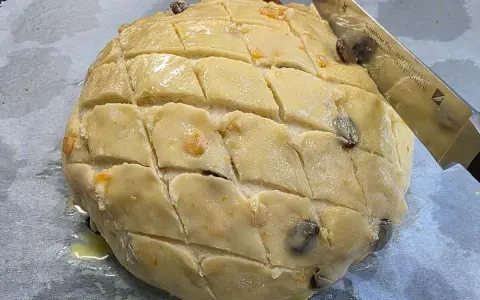 Ein runder Teigling mit eingeschnittenen Rauten, bereit für das Osterfladen Rezept. Rosinen und Orangenschale sind sichtbar.