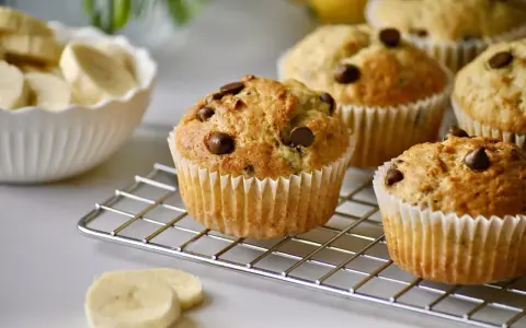 Frisch gebackene schoko bananen muffins mit Schokostückchen auf einem Gitter. Daneben eine Schüssel mit Bananenscheiben.