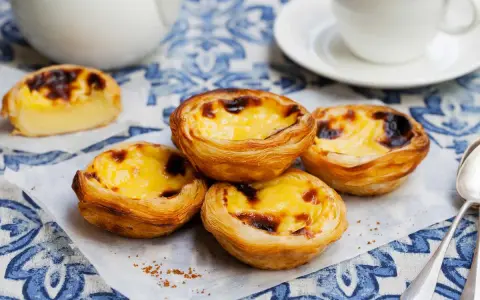 Vier köstliche Pastel de Nata auf einem blauen Tischtuch, mit einer Tasse Kaffee im Hintergrund.