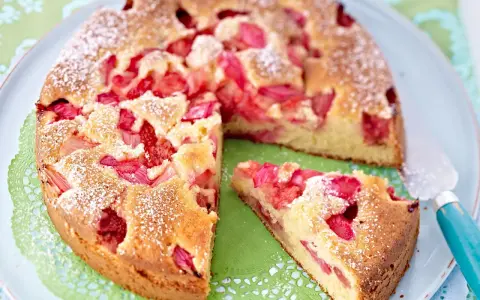 Ein saftiger Erdbeer Rhabarber Kuchen, bestäubt mit Puderzucker, liegt auf einem Teller. Ein Stück wurde bereits entnommen.
