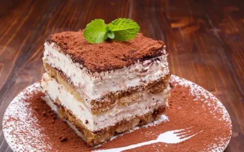 Ein Stück cremiges Tiramisu ohne Ei, bestäubt mit Kakao und garniert mit Minze, auf einem Teller mit Kakaopulver und einer Gabel.