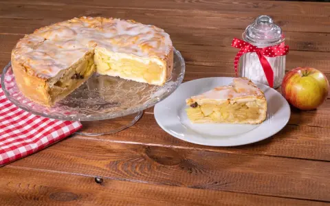 Omas weltbester Apfelkuchen, saftig und mit Zuckerguss überzogen, wartet auf einem Tortenständer. Ein Stück auf einem Teller und ein Apfel daneben laden zum Genießen ein.