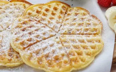 Zuckerbestäubte Buttermilchwaffeln, die auf einem Holzbrett liegen. Daneben sind Himbeeren und Bananenscheiben zu sehen.