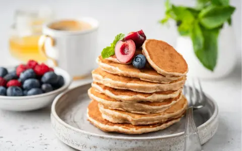 Ein Stapel veganer Pancakes, garniert mit Kirschen und Blaubeeren, daneben eine Schale Beeren und eine Tasse Kaffee.
