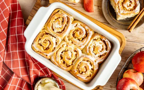 Frisch gebackene Apfel Zimt Schnecken mit Zuckerguss in einer Auflaufform. Daneben Äpfel und Zimtstangen.