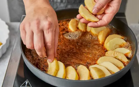 Hände legen Apfelspalten in eine Pfanne mit karamellisierter Butter. Die Zubereitung der klassischen Tarte Tatin beginnt.
