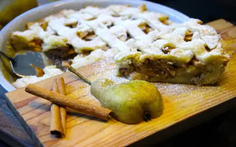 Ein köstlicher Birnenkuchen mit Gittermuster, bestäubt mit Puderzucker. Daneben liegen eine Birne und Zimtstangen – perfekt für ein Birnenkuchen Rezept.