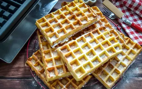 Frisch gebackene belgische Waffeln, mit Puderzucker bestäubt, liegen auf einem Gitter. Daneben ein Waffeleisen und eine karierte Serviette.