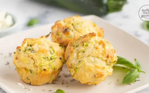 Drei goldbraune Zucchini-Muffins mit Kräutern und Käse auf einem Teller, garniert mit Basilikum.