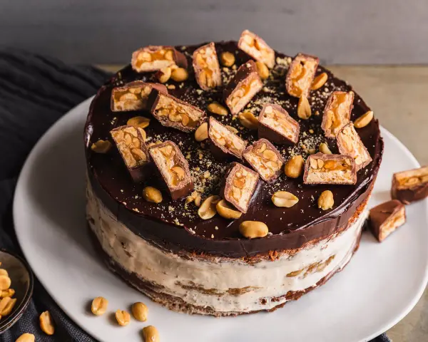 Eine köstliche Snickers Torte mit Schokoladenglasur, Erdnüssen und vielen Snickers-Stücken obendrauf.