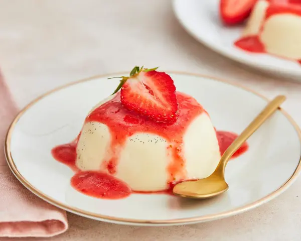 Cremige Panna Cotta mit Erdbeersauce und frischer Erdbeere, garniert mit Vanillepunkten. Eine goldene Löffel liegt daneben.