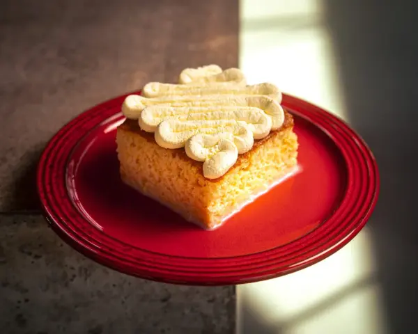 Ein Stück saftiger Tres Leches Kuchen mit Sahnetupfern auf einem roten Teller, bereit zum Genießen.