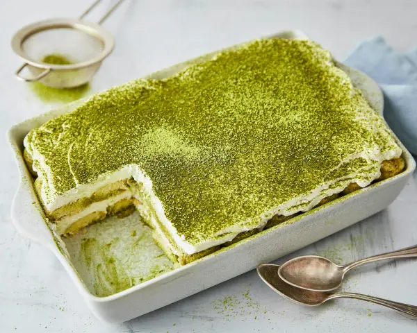 Ein cremiges Matcha Tiramisu, bestäubt mit feinem grünen Pulver, wartet in einer Auflaufform darauf, genossen zu werden. Löffel liegen bereit.