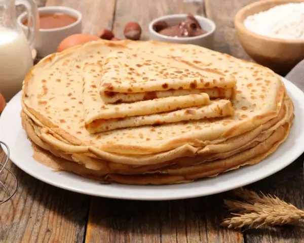 Ein Stapel goldbrauner Pfannkuchen, zubereitet nach einem sehr guten Pfannkuchen Rezept, daneben Zutaten wie Milch, Eier, Mehl, Marmelade und Nuss-Nougat-Creme.