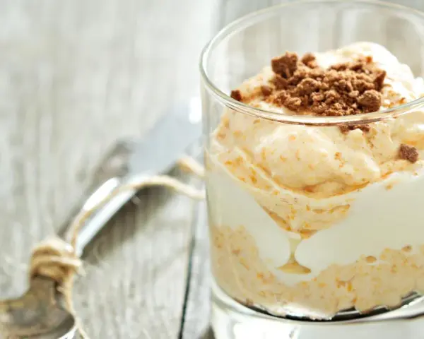 Cremiger cheesecake im glas, geschichtet mit weißer Creme und garniert mit zerbröselten Keksen. Ein Löffel liegt daneben.