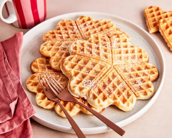 Herzförmige Waffeln, bestäubt mit Puderzucker. Ein köstlicher waffelteig ohne butter, serviert auf einem Teller mit zwei Gabeln.