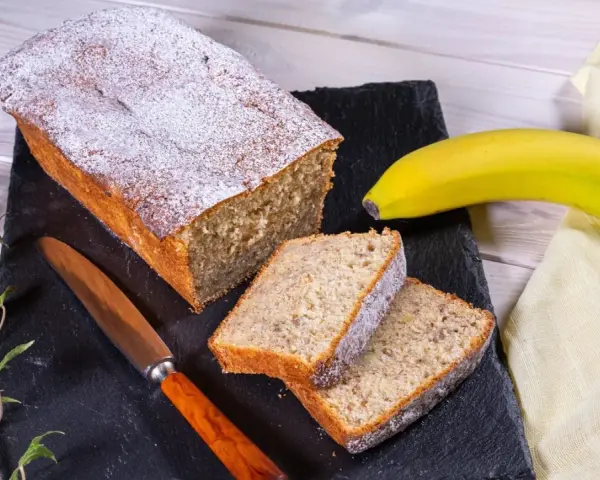 Saftiger Bananenkuchen, bestäubt mit Puderzucker, neben einer Banane und einem Messer. Perfekt für ein Bananenkuchen Rezept.