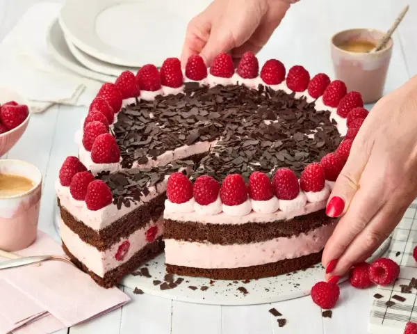 Eine köstliche Schoko Himbeer Torte, verziert mit frischen Himbeeren und Schokostreuseln, wird von Händen arrangiert.