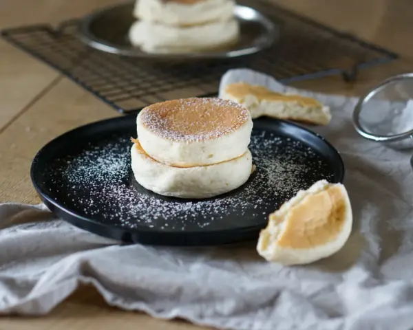 Zwei fluffige japanische Pancakes auf einem schwarzen Teller, bestäubt mit Puderzucker. Daneben ein Stück Pancake und ein Sieb.