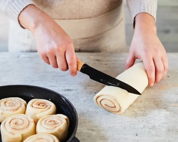 Hände schneiden Teigrolle für Zimtschnecken. Bereits geformte Schnecken warten in einer Pfanne auf das Backen. Ein tolles Zimtschnecken Rezept!
